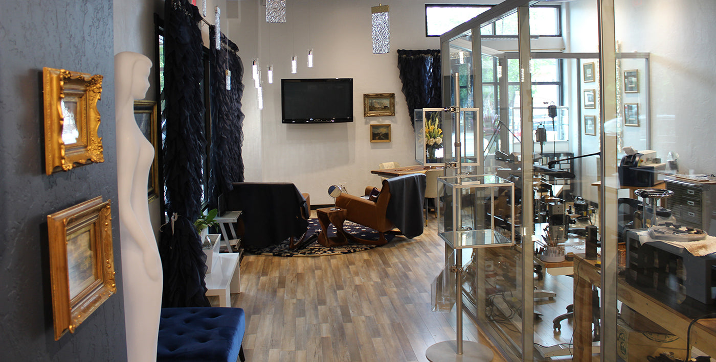 A boutique interior with a glass display case on the right, a mannequin torso and framed gold frames on the left, and a furnished room with a chair, table, TV, and wood flooring in the background.
