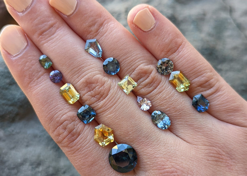 A close-up of a hand displaying multiple loose gemstones in various shapes and colors, including blue, yellow, purple, and dark gray, set against a natural gray rock background.