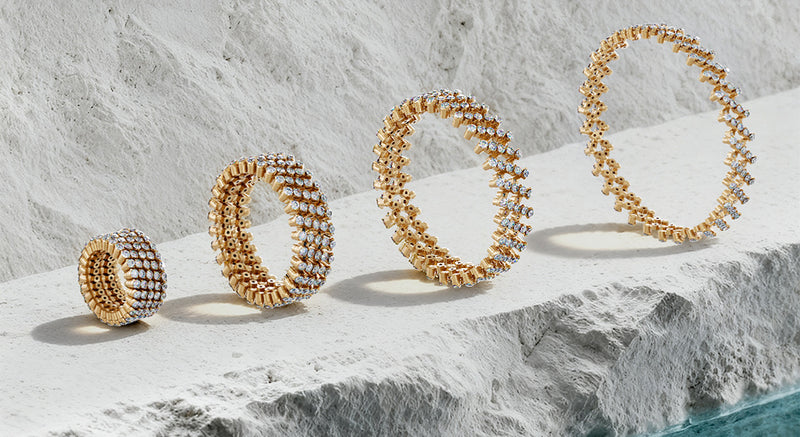 Four gold-toned beaded rings of varying sizes arranged diagonally on a textured white stone surface.